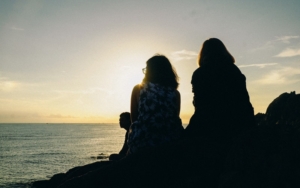 two women sitting side by side looking out over the water with sun setting