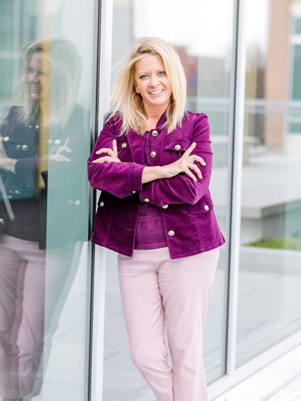 Sherrie leaning against a glass wall with her arms crossed, smiling