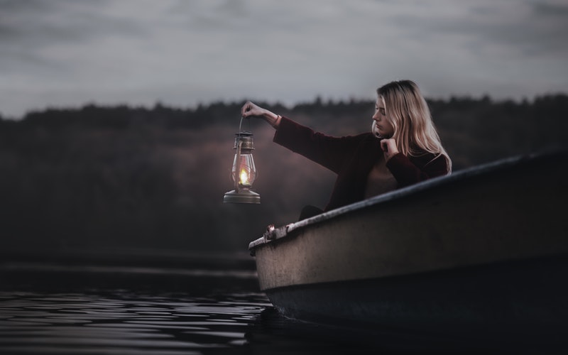 woman in a boat on a lake, blond hair, dark colored sweater, in the dusk of the day and is holding a kerosene lamp over the side of the boat.