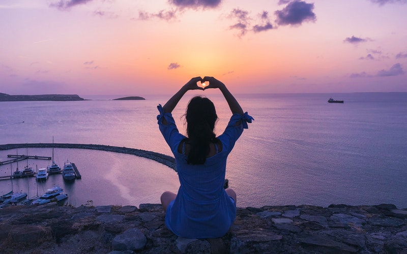 defender, woman sitting on rocks overlooking water facing the sunset, arms raised overhead forming the heart symbol with her hands placed perfectly around the sun in the horizon