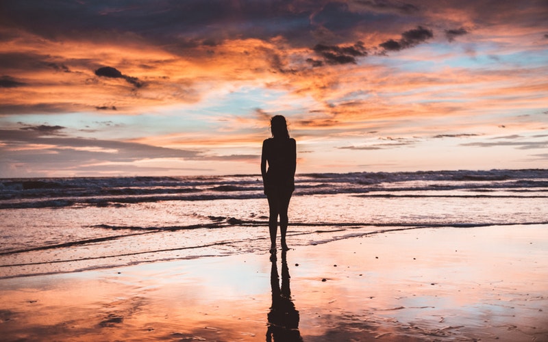 highs and lows, shilouette of woman facing ocean, sky is dreamsicle orange and blue clouds reflected on the wet sand