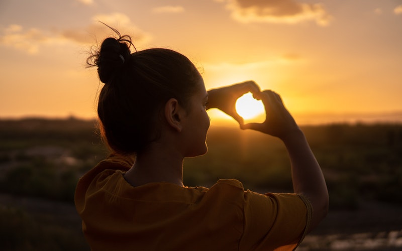 His Love, sunset, woman facing the sun with hands in front of her face, making the heart symbol around the sun that is off in the distance.