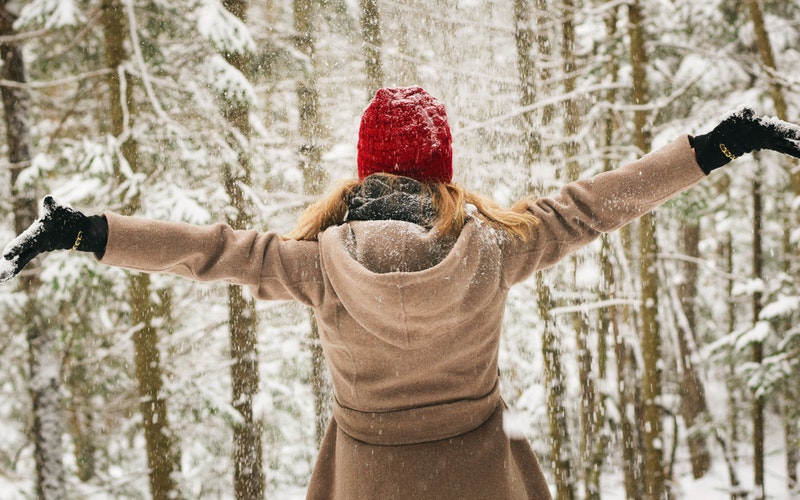 Joyful Noise, trees covered in snow, woman with snow dusting on her, red hat, camel colored coat and black gloves. She's facing away and has both her arms extended on either side of her.