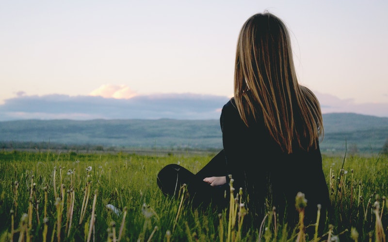 woman facing away, sitting in a grass field staring out into the distant mountains