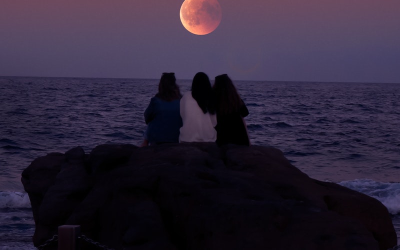 Three women sitting on a rock looking at the moon rise over the ocean, For Your Good and His Glory