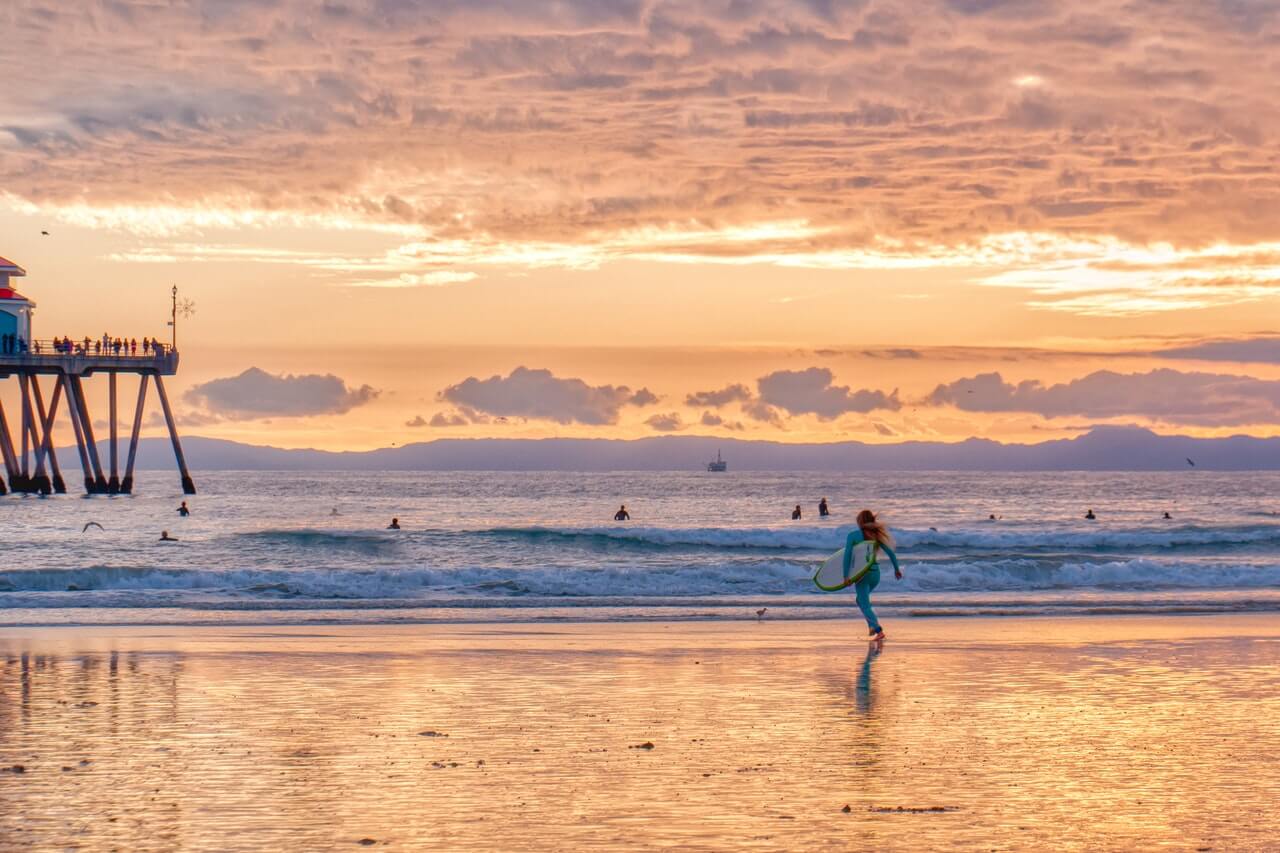 Life Verse - Girl running into the surf at sunset with her surfboard under her arm. Pier is off to the left with people on the railing. Surfers in the water
