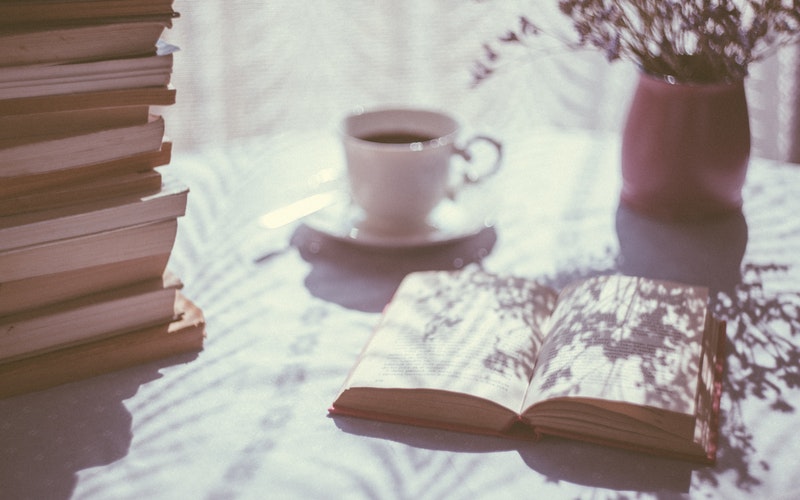 table against a window with light coming through. Books stacked to the left, the Word (book) opened in the middle with a cup of tea and a vase of flowers
