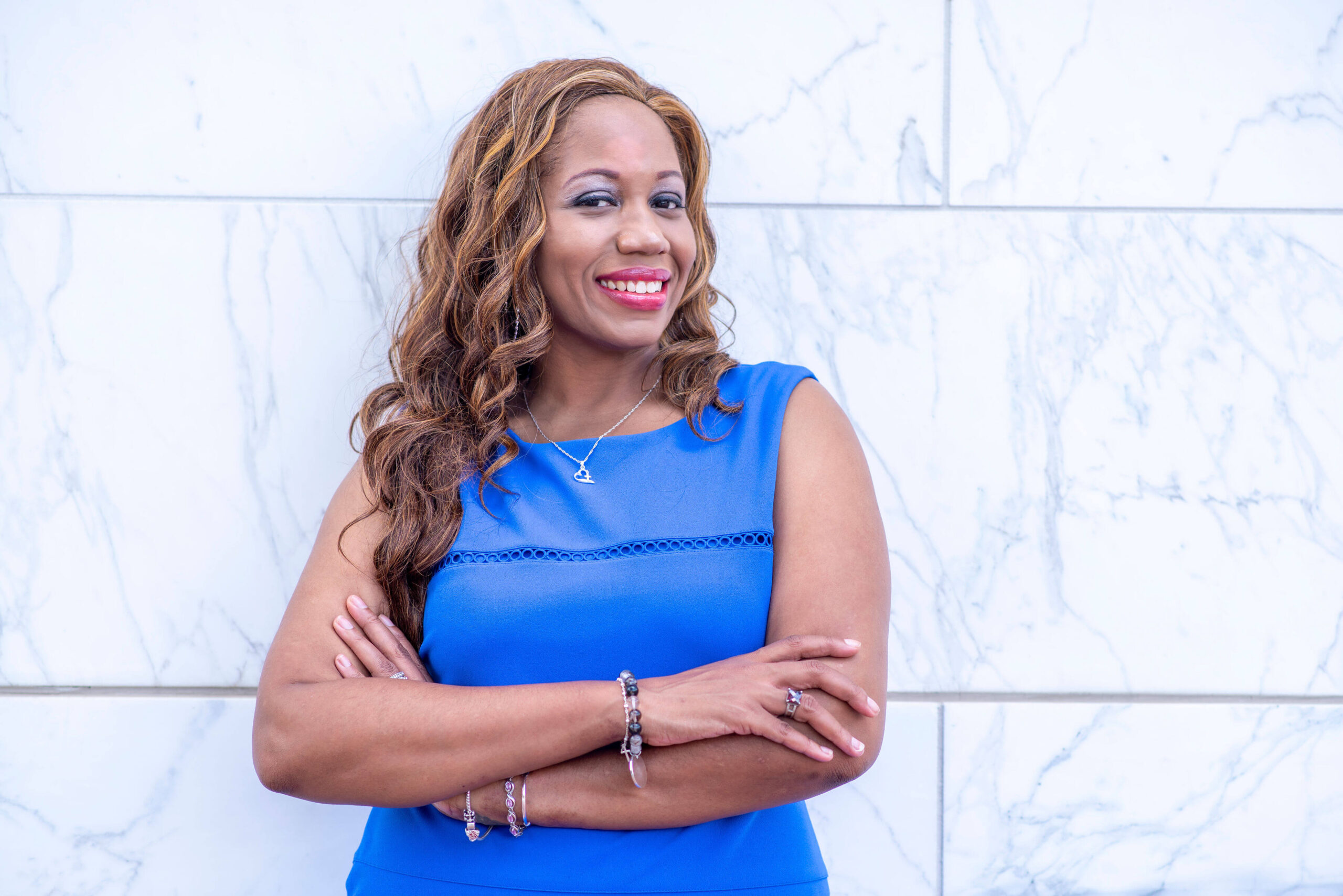 headshot of Life Coach, Artisha Bolding, an African American with long hair, standing against a white marble wall wearing a blue dress